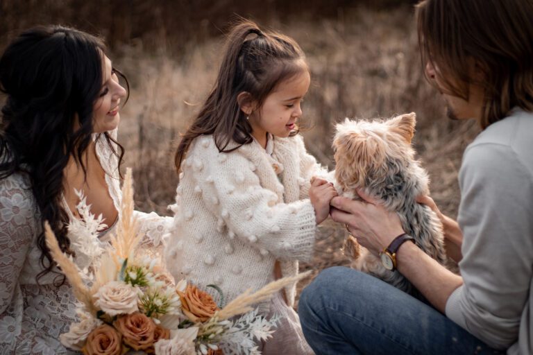 child, dad, and pregnant mom in ivory flowy dress playing with dog in long grass field by elk lake for maternity photoshoot victoria bc with alyssa orrego photography